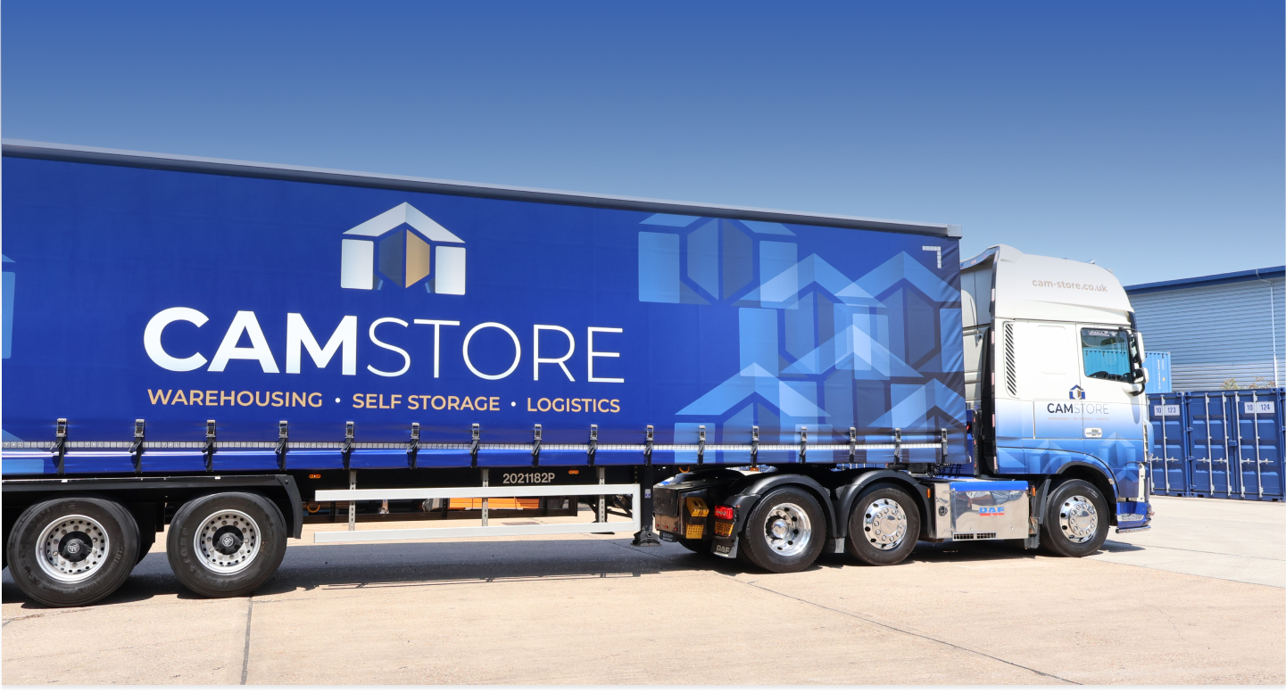 Blue lorry with black wheels and white cabin front. Lorry has big CAMSTORE logo on it. Self-storage containers behind.